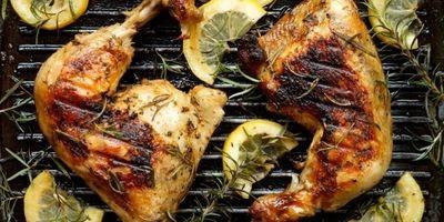 Two charred chicken legs on a grill garnished with lemon slices and fresh rosemary